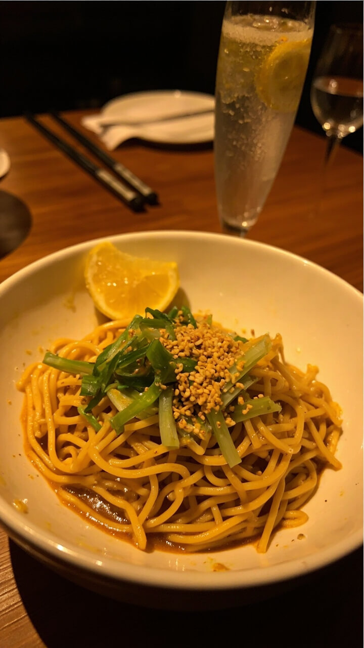 A wide white soup bowl filled with glistening ramen noodles coated in golden butter sauce, sliced scallions, and a sprinkle of toasted sesame seeds on top. A wedge of lemon sits on the rim. The table is set with chopsticks and a small glass of sparkling water.