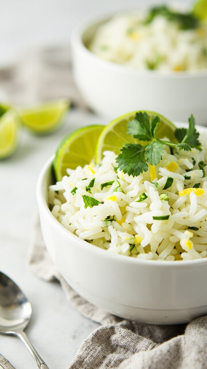 A white serving bowl filled with fluffy, white rice speckled with finely chopped fresh cilantro and flecks of grated lime zest. Lime wedges are arranged on one side with a sprig of cilantro on top. Next to the bowl sits a folded kitchen towel and a spoon. A white serving bowl filled with fluffy, white rice speckled with finely chopped fresh cilantro and flecks of grated lime zest. Lime wedges are arranged on one side with a sprig of cilantro on top. Next to the bowl sits a folded kitchen towel and a spoon.
