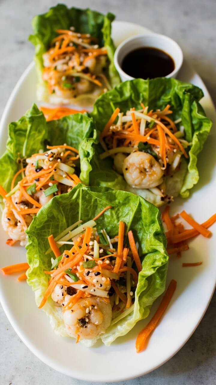 Crisp romaine leaves filled with sautéed ginger shrimp, thin carrot ribbons, green onion slices, and a sprinkle of sesame seeds. The cups are nestled on a white oval plate with little dipping bowls of soy sauce and a few extra carrot shreds on the side.