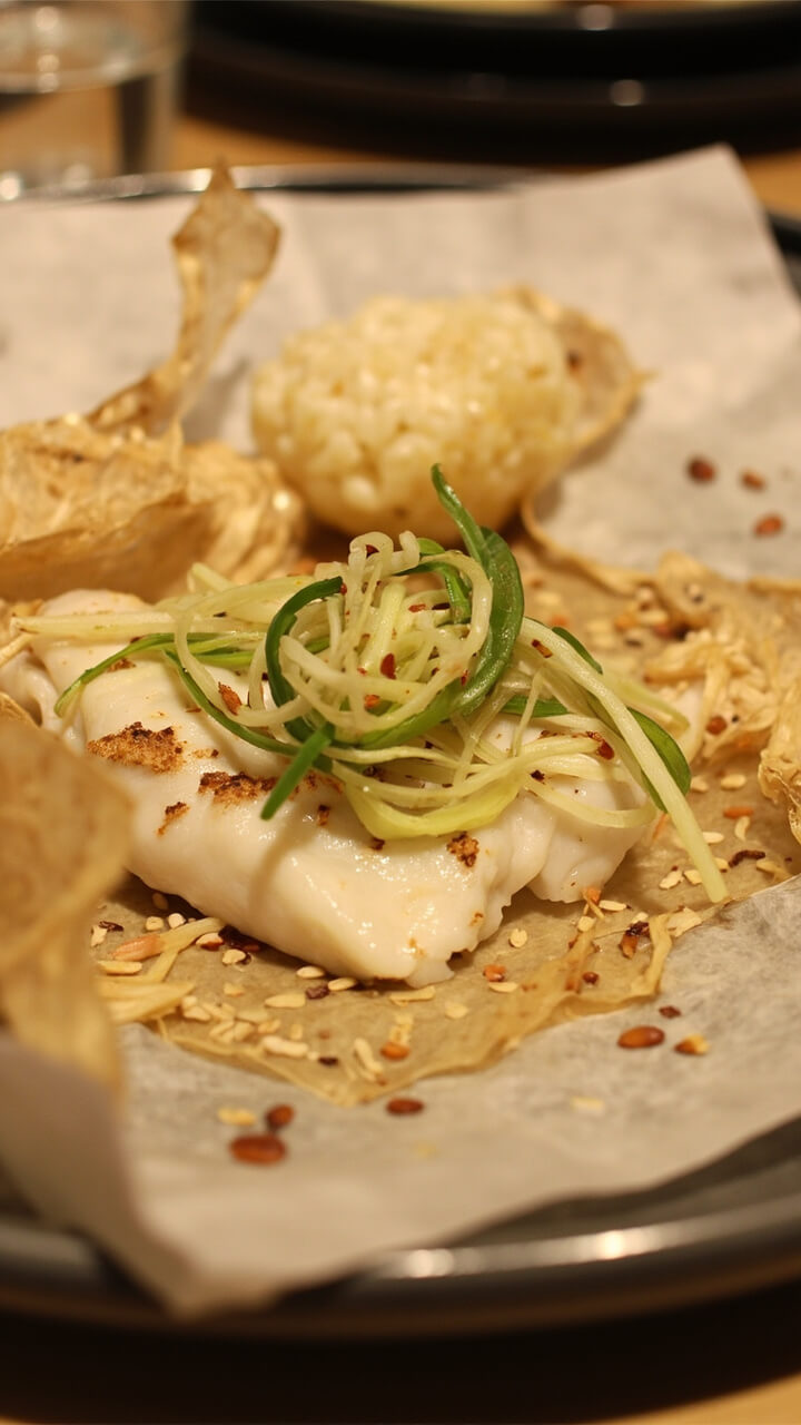 A plate with a delicate white fish fillet nested in crinkled parchment, topped with julienned ginger and scallions, edges flecked with sesame seeds. Some jasmine rice on the side. A plate with a delicate white fish fillet nested in crinkled parchment, topped with julienned ginger and scallions, edges flecked with sesame seeds. Some jasmine rice on the side.