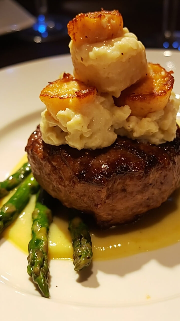 A thick filet mignon on a white plate topped with lump crab meat and two golden scallops, all drizzled with buttery béarnaise sauce. Asparagus spears are artfully arranged around the steak. A thick filet mignon on a white plate topped with lump crab meat and two golden scallops, all drizzled with buttery béarnaise sauce. Asparagus spears are artfully arranged around the steak.