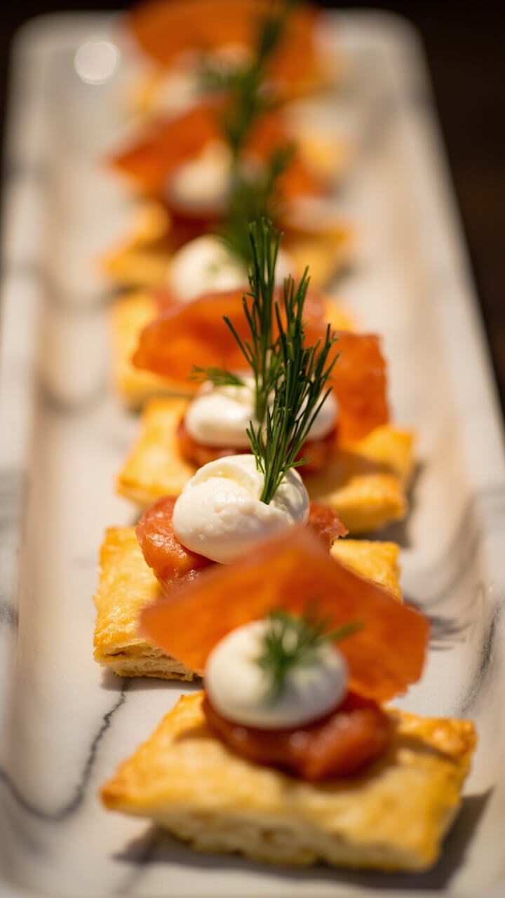 Small golden puff pastry squares topped with a swirl of cream cheese, a sliver of smoked salmon, and a sprig of fresh dill arranged on a marble platter.