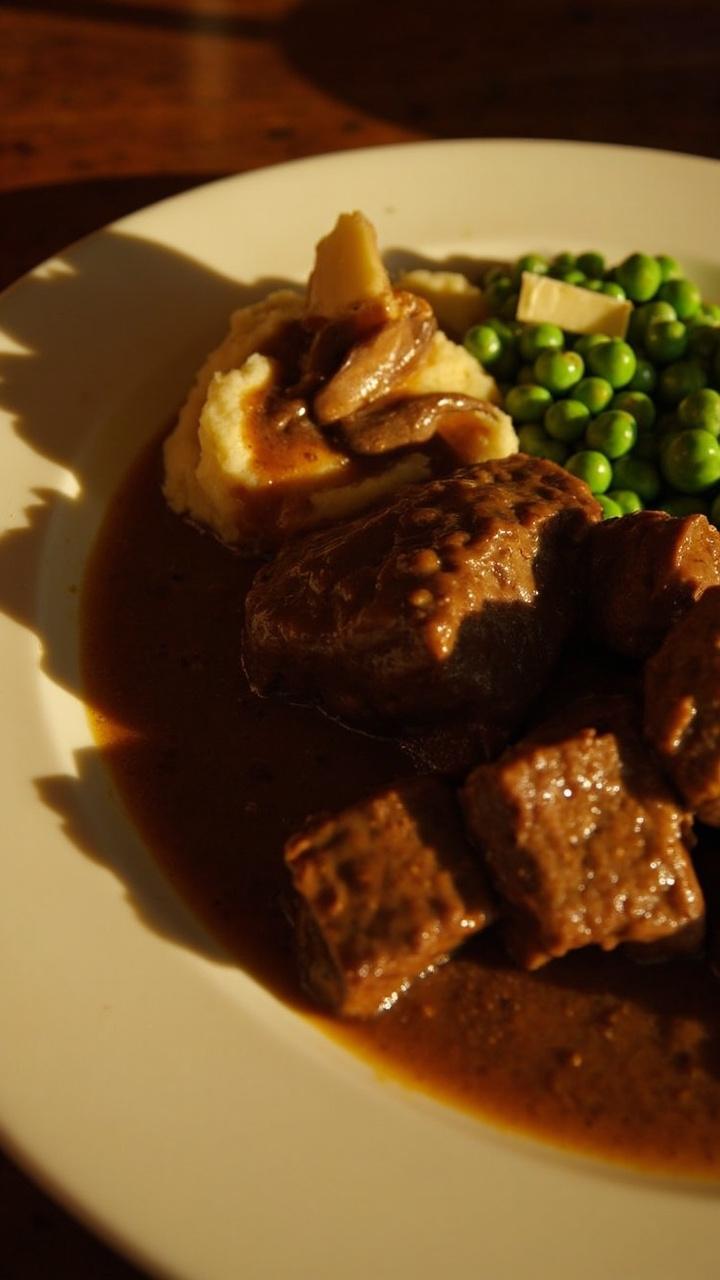 Cubed steaks in mushroom-brown gravy, served in a white rimmed plate with mashed potatoes swirled beside and a side of buttered green peas.