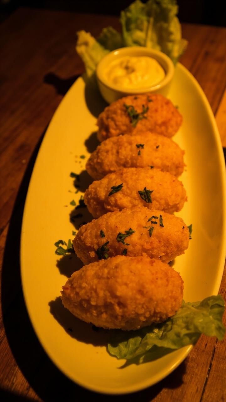 Six oval-shaped croquettes with a crisp, golden-brown coating, arranged in a tidy row on a white oval platter. Each is garnished with chopped parsley, with a small bowl of creamy dip to the side and a few crisp lettuce leaves on the platter.