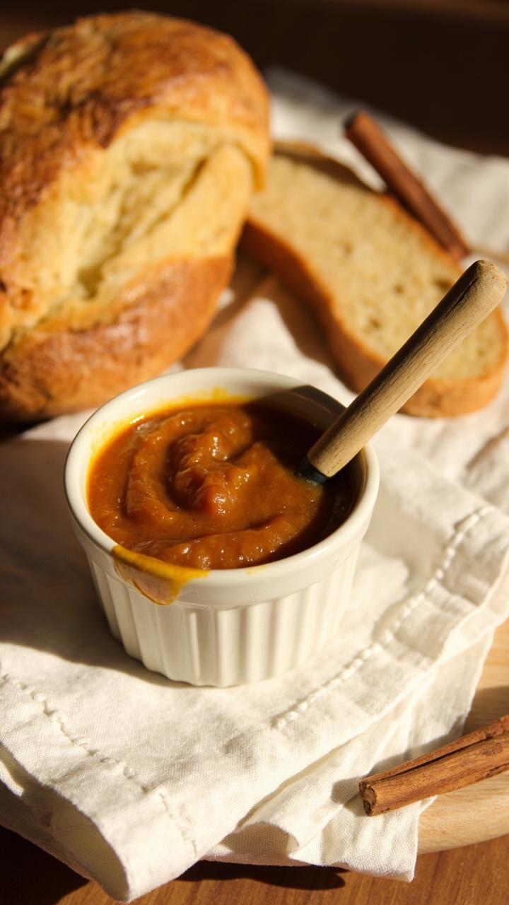 A small ceramic ramekin filled with deep amber crab apple butter, with a wooden spoon sunk in. Thick slices of sourdough and cinnamon sticks are arranged alongside on a linen napkin.