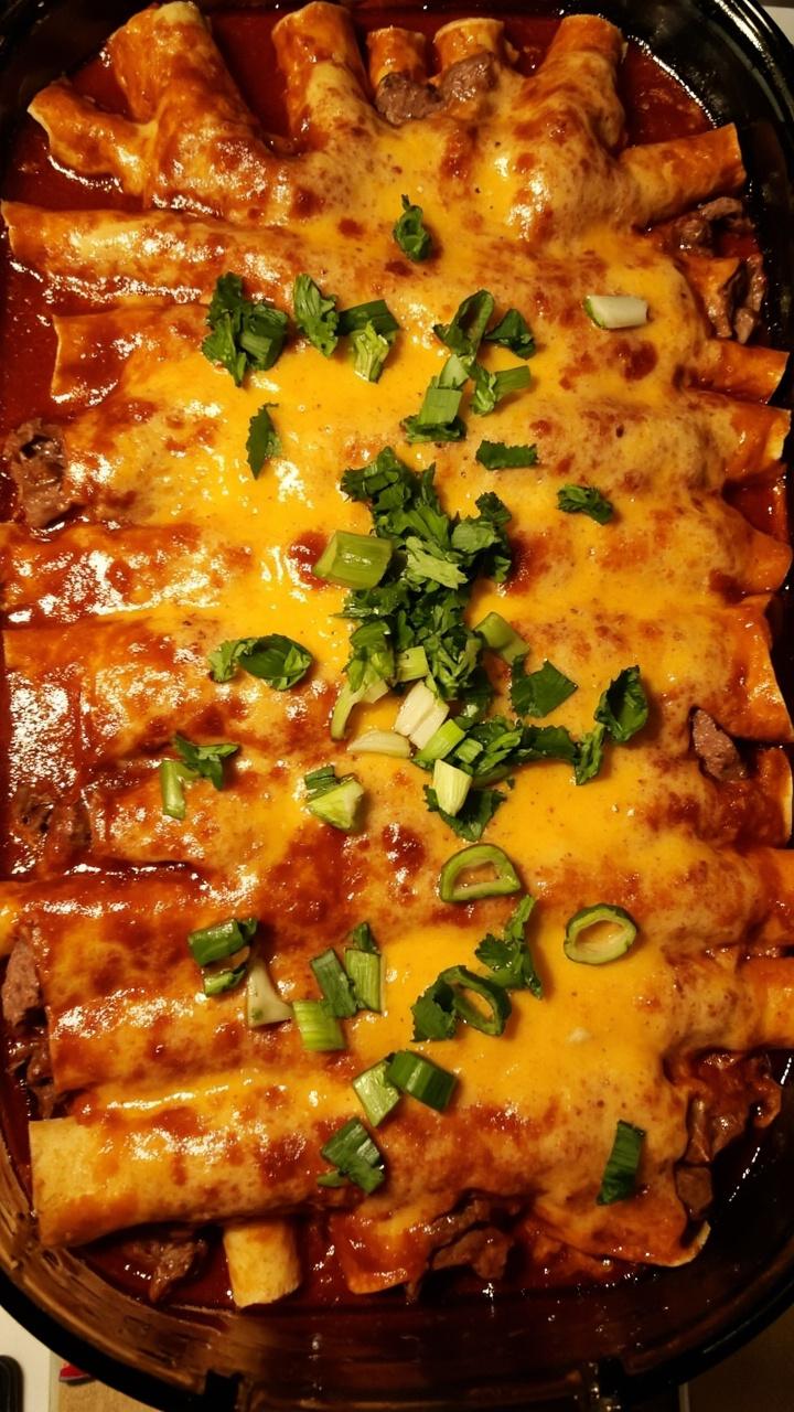 A baking dish filled with rolled tortillas covered in red enchilada sauce and bubbling melted cheese. Chopped steak peeks out from the rolled ends, and the tray is sprinkled with chopped green onions and cilantro.