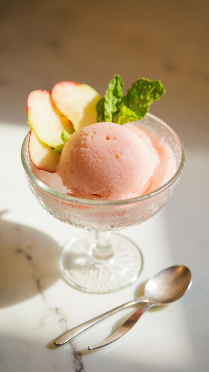 A small glass dish of smooth, blush-pink sorbet with a few thin crab apple slices and a sprig of fresh mint, set on a marble countertop with frosted spoons.