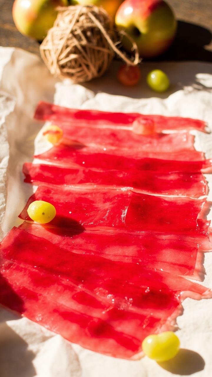 A sheet of translucent, rosy-red fruit leather cut into neat, rollable strips, arrayed on white parchment. Small crab apples and a ball of twine for rolling are set beside the strips.