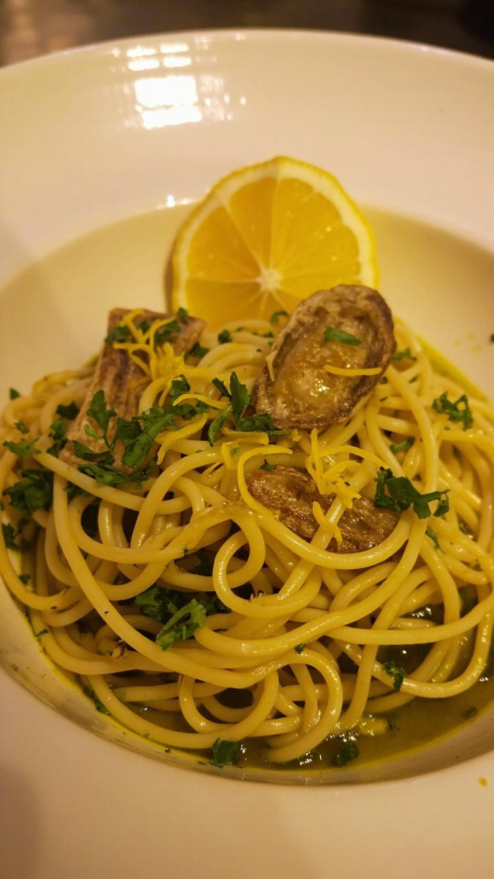 A shallow pasta bowl with spaghetti tossed in a glossy olive oil sauce, flecks of parsley and lemon zest on top, and a few anchovy fillets as garnish. A wedge of lemon rests on the rim.