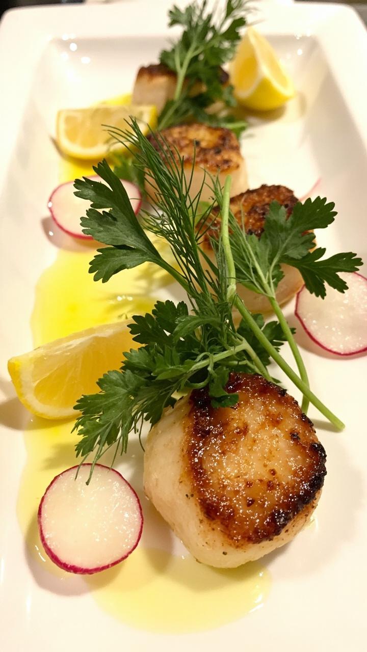 Golden-brown scallops arranged on a small white plate, topped with a tangle of fresh parsley, dill, and thinly sliced radishes. Lemon wedges on the side, with a light drizzle of olive oil.