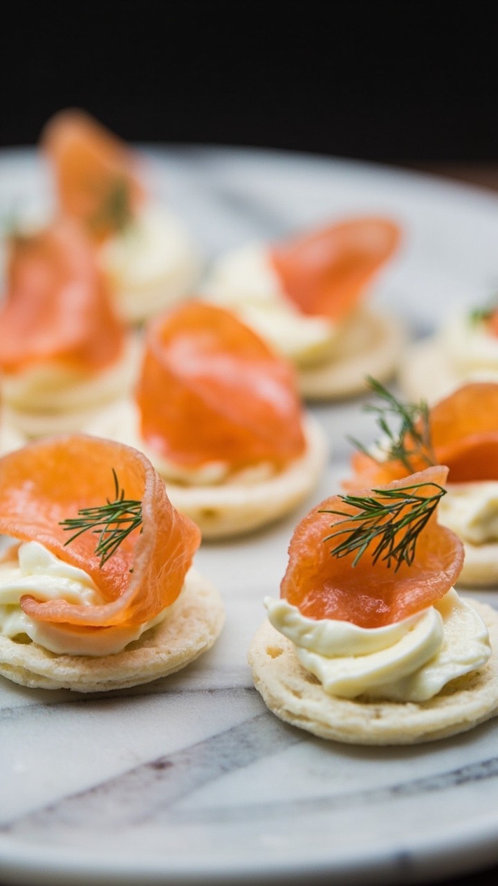 Pale, fluffy silver-dollar blini topped with a curl of smoked salmon, a small swirl of lemon crème, and a pinch of chopped dill, arranged neatly on a marble serving slab. Pale, fluffy silver-dollar blini topped with a curl of smoked salmon, a small swirl of lemon crème, and a pinch of chopped dill, arranged neatly on a marble serving slab.