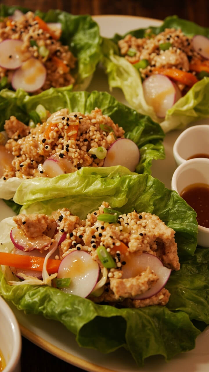 A platter with crisp butter lettuce leaves, each filled with a mixture of crab, thinly sliced vegetables, and a drizzle of sesame dressing. There’s a sprinkle of sesame seeds and slivers of green onion. Small dipping bowls are on the side. A platter with crisp butter lettuce leaves, each filled with a mixture of crab, thinly sliced vegetables, and a drizzle of sesame dressing. There’s a sprinkle of sesame seeds and slivers of green onion. Small dipping bowls are on the side.