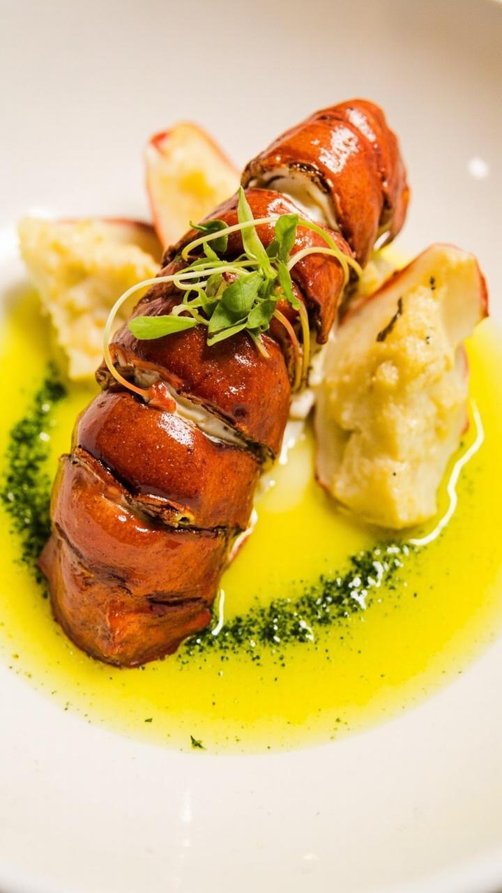 A peeled lobster tail sits in the center of a shallow, white bowl, drenched in glistening yellow butter. Claw meat is arranged delicately beside it. Brilliant green chive oil is drizzled in a ring around the lobster, and a few microgreens rest atop for garnish.