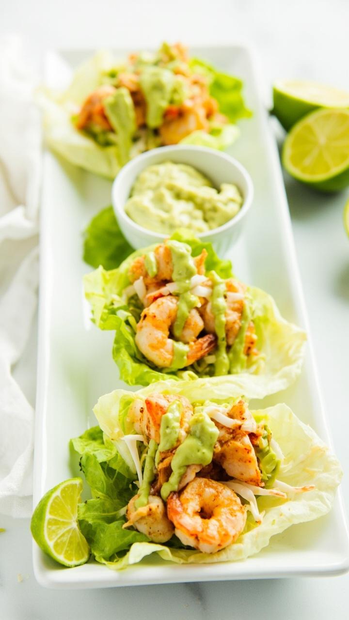 Three butter lettuce cups lined up on a long white platter each filled with lime kissed shrimp shredded cabbage and a drizzle of avocado crema. Lime halves to squeeze on the side. A small bowl of extra crema nearby.