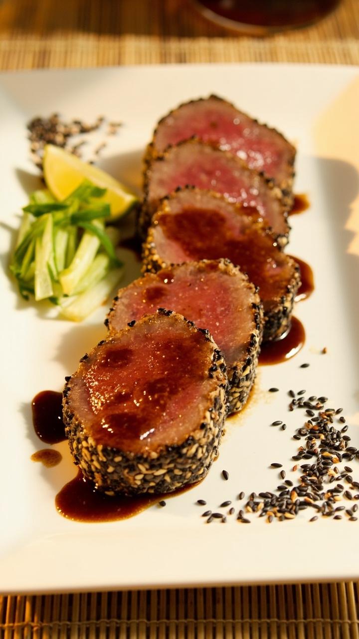 Four sesame crusted tuna steaks seared to rare in the center, sliced into thick medallions and fanned on a long white plate, a small drizzle of glossy soy ginger glaze over the top, thinly sliced scallions and a lime wedge, a scattering of black and white sesame seeds, a bamboo placemat under the plate