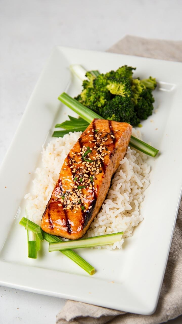 A rectangular white plate with a fillet of glazed teriyaki salmon resting on a neat mound of steamed rice, sprinkled with sesame seeds and thinly sliced scallions, a small pile of steamed broccoli on the side.