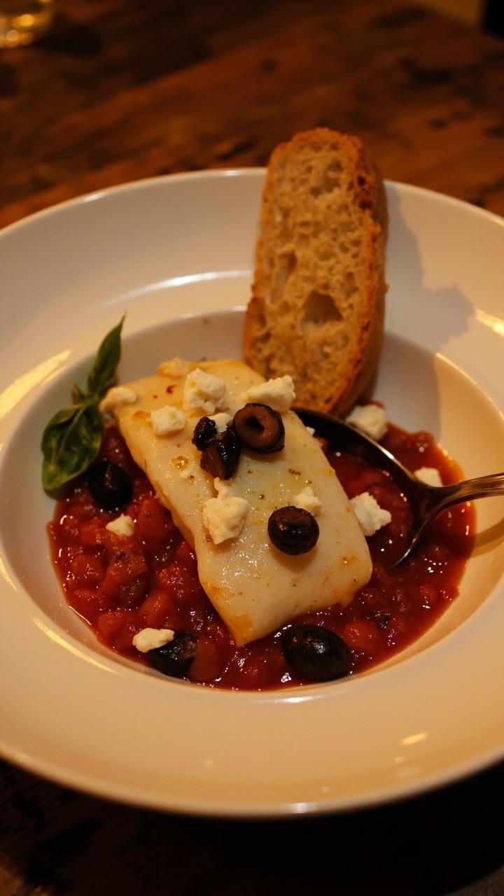 A shallow white bowl with a ladle of bright tomato braise. One cod fillet nestled in the sauce topped with chopped olives and crumbled feta. A slice of crusty bread leaning on the rim and a sprig of basil.