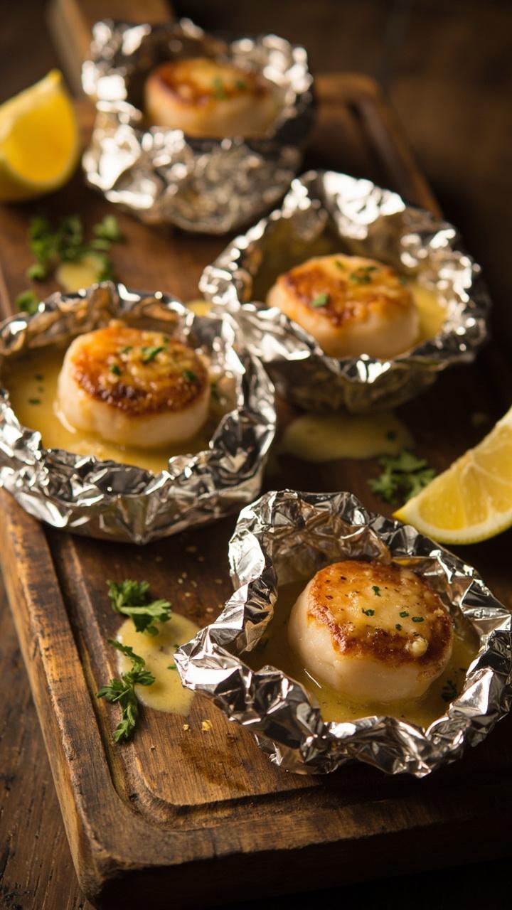 Individual foil packets unwrapped on a rustic wooden board revealing scallops with melted Parmesan, small pools of butter and garlic, a sprinkling of parsley, a wedge of lemon beside each packet.
