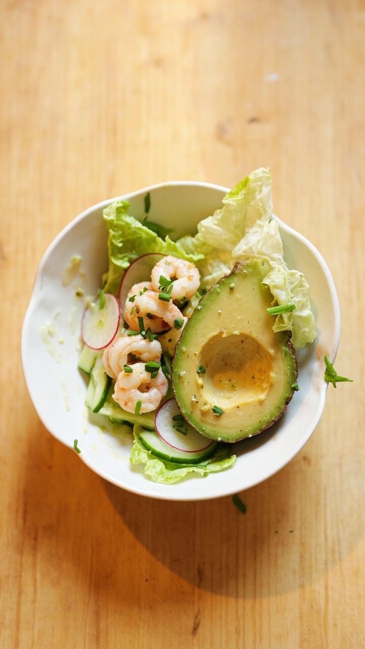 A shallow bowl with halved avocado, a small pile of butter lettuce, sliced cucumber, radish rounds, and four to five poached shrimp arranged on top. A drizzle of pale vinaigrette and a scattering of chopped chives. A light wooden table surface. A shallow bowl with halved avocado, a small pile of butter lettuce, sliced cucumber, radish rounds, and four to five poached shrimp arranged on top. A drizzle of pale vinaigrette and a scattering of chopped chives. A light wooden table surface.