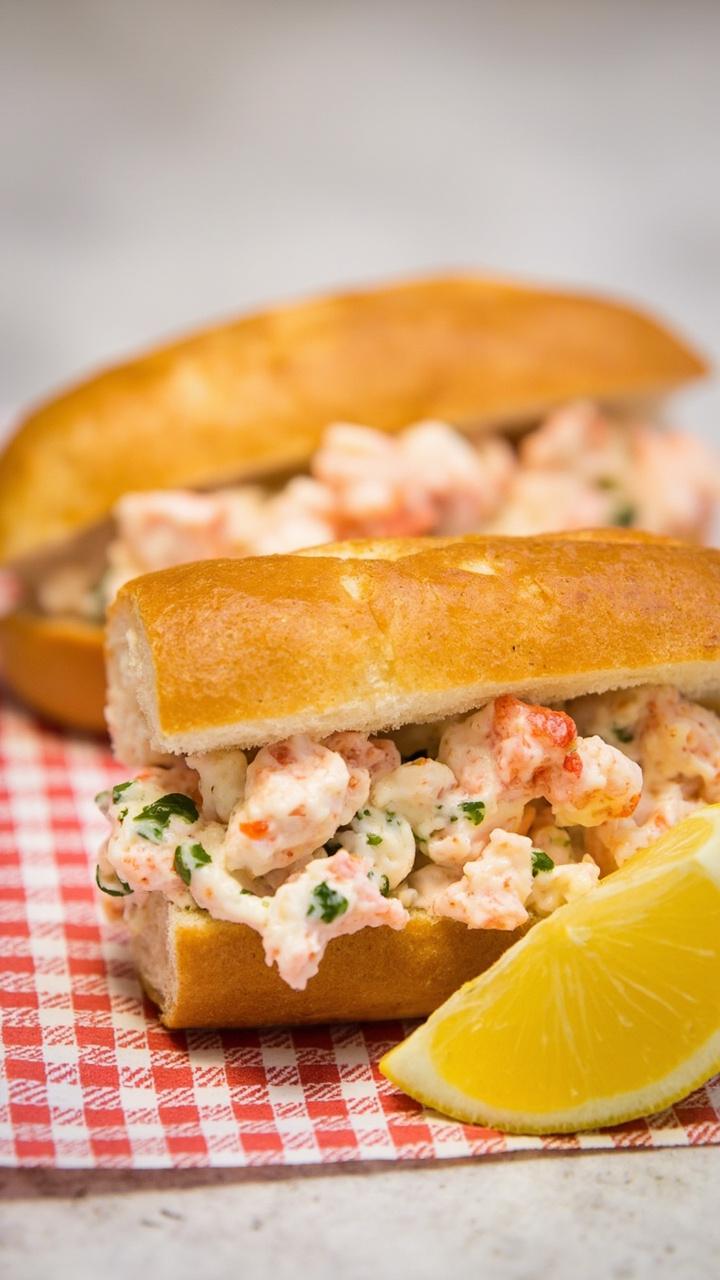 Two toasted split top rolls buttered and golden, generous lobster filling peeking out with flecks of green herb mayo, lemon wedge on the side, checked napkin underneath Two toasted split top rolls buttered and golden, generous lobster filling peeking out with flecks of green herb mayo, lemon wedge on the side, checked napkin underneath
