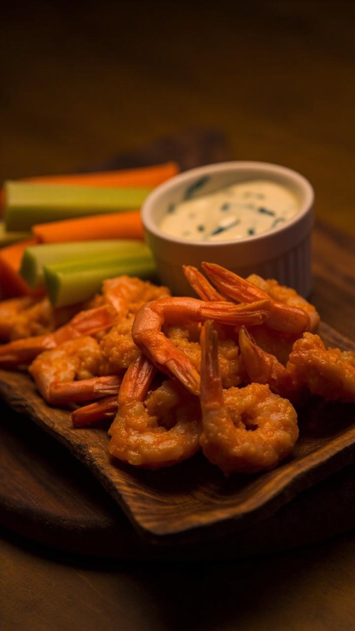 orange sauced shrimp piled on a small platter, celery sticks and carrot sticks arranged beside them, a small ramekin of blue cheese dressing, dark wood board