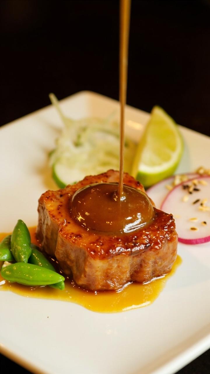 A seared tuna steak glazed with a glossy amber miso sauce on a small square plate, a few steamed edamame pods and thinly sliced radish, sesame seeds and a small lime wedge